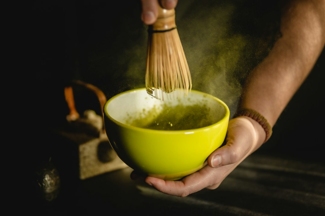 Persona preparando matcha ceremonial con batidor de bambú en tazón amarillo sobre fondo oscuro