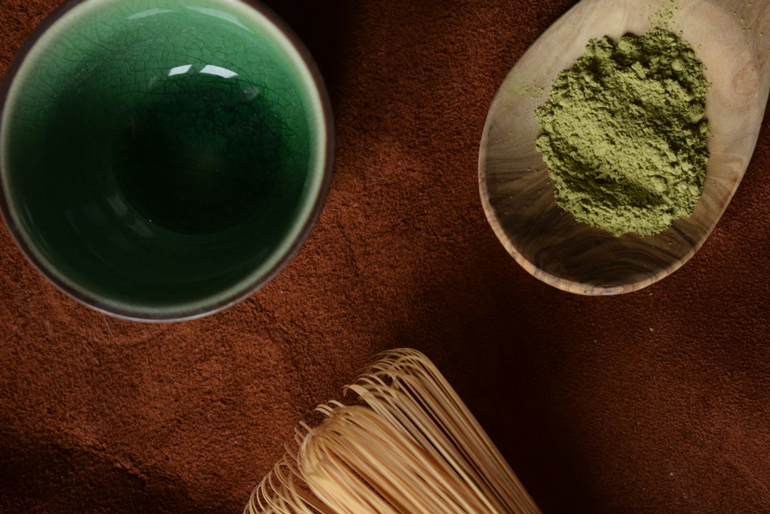 Cuenco de cerámica verde con matcha ceremonial líquido, cucharilla de bambú y polvo de matcha en recipiente de madera