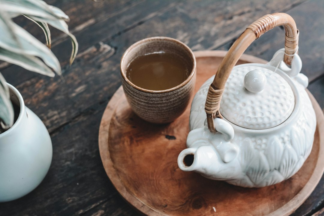 Taza de cerámica con té sobre bandeja de madera, tetera blanca y planta verde en mesa oscura