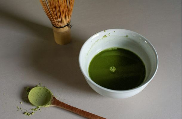Polvo de matcha verde en cucharilla de madera, batidor de bambú y tazón blanco con matcha ceremonial preparado