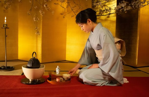 Mujer en kimono gris realizando ceremonia del té japonesa sentada sobre tatami rojo con tetera y utensilios tradicionales