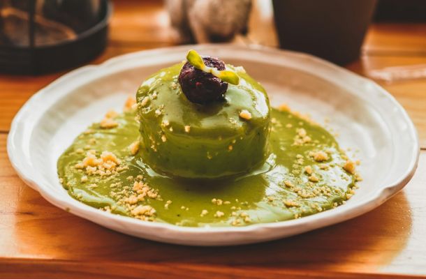 Postre gourmet con crema verde de matcha, salsa de matcha, granola dorada y flor comestible sobre plato blanco