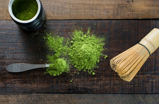 Polvo de matcha ceremonial verde esparcido sobre tabla de madera con batidor de bambú, cuchara medidora y frasco de